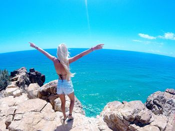 Rear view of woman with arms outstretched standing on cliff by sea against blue sky