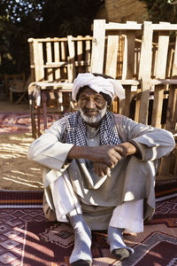 Aged nubian man in ethnic outfit sitting on carpet in yard and looking at camera in sunlight