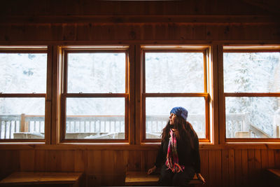 Woman in outerwear sitting on bench near windows and looking away inside lumber hut on winter day in valley of the ghosts in monts valin national park in quebec, canada