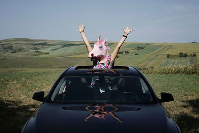 Woman with arms raised at sunroof of car while wearing unicorn mask