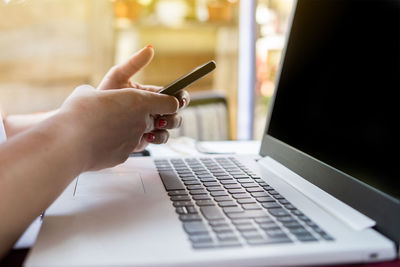 Cropped hand of woman using mobile phone over laptop