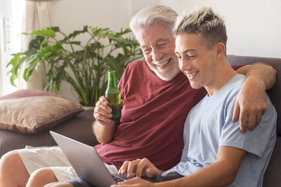 Smiling grandfather with grandson using laptop while holding beer sitting at home