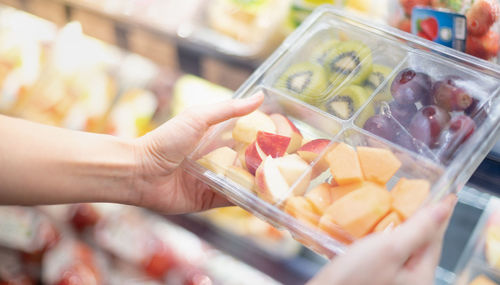 Female hand holding mix of fresh fruit in the transparent plastic box with a fork in a supermarket.