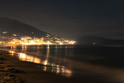 Illuminated city by sea against sky at night