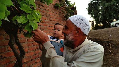 Smiling grandfather with grandson looking at plant while standing against brick wall