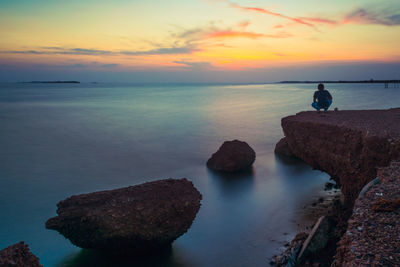 Rear view of man crouching on cliff by sea against dramatic sky during sunset