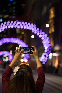 Rear view of woman photographing illuminated lights from mobile phone at night
