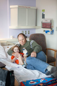 Worried father snuggles with silly toddler in hospital bed.