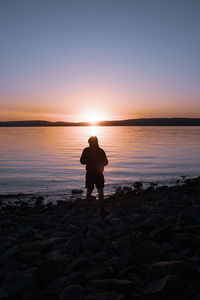 Man standing on rock at lakeshore against sky during sunset