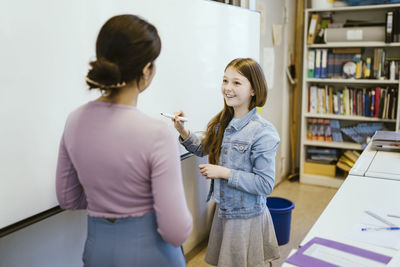 Smiling female student talking to teacher standing near whiteboard in classroom