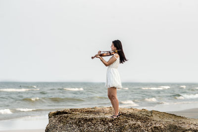 Young woman playing violin while standing on rock at beach