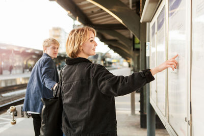 Young man pulling teenage girl pointing on information board at railroad station platform