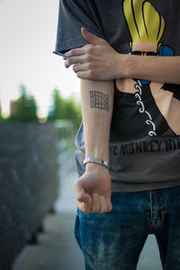 Midsection of young man showing tattoo while standing on street against sky