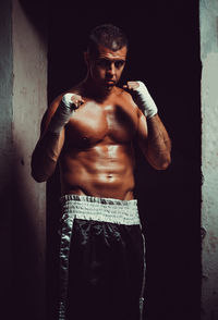 Portrait of shirtless male athlete wearing hand wrap while standing against wall