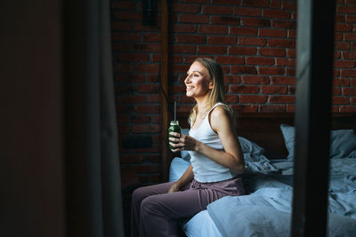 Young adult woman with blonde long hair in casual with green smoothie near window at the home