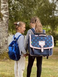 Rear view of girls sisters standing in park with backpacks