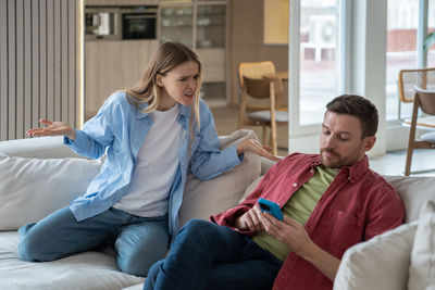 Family quarrel. focused man hang out on smartphone ignoring unpleased woman sitting on couch at home