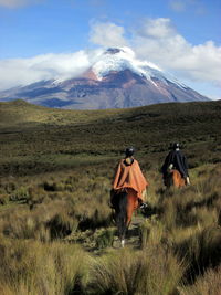 Rear view of people riding horses on grassy landscape against cotopaxi