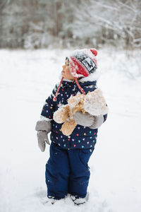 Toddler child standing outside in winter scenery holding soft toy.