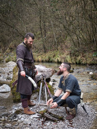 Men wearing costume by stream igniting firewood in forest