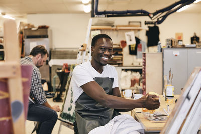 Portrait of happy young male student in apron sitting at table in art class