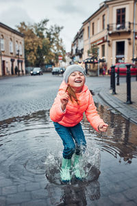 Happy smiling girl jumping in the puddle during walk in a downtown on rainy gloomy autumn day