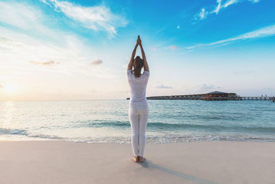 Rear view of woman with arms raised meditating while standing on shore at beach against sky during sunset