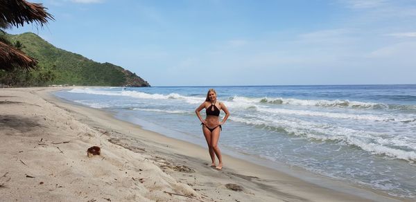 Full length of woman in bikini standing on shore at beach against sky