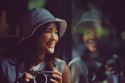 Smiling woman holding camera while leaning on glass window