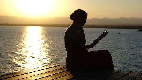 Silhouette woman reading on pier over lake against sky during sunset