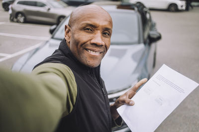 Smiling bald man showing car contract while taking selfie