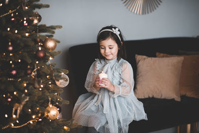 Cute girl holding candle sitting on sofa by illuminated christmas tree