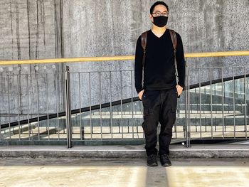 Full length of young asian man standing against railings, travelator and textured wall.