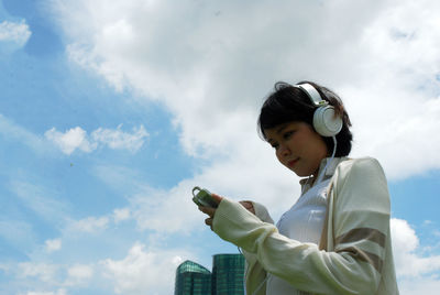 Low angle view of woman listening music while using smart phone against sky