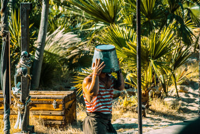 Mid adult man with bucket on head standing against palm trees