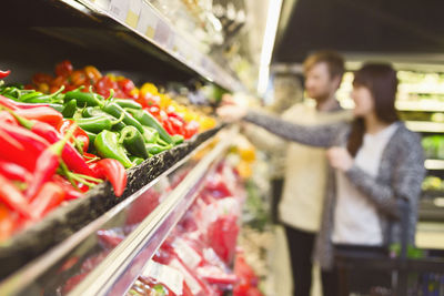 Chili peppers arranged in shelf with couple shopping in background at supermarket