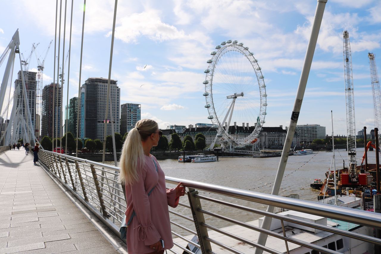 sky, one person, real people, architecture, built structure, connection, building exterior, lifestyles, railing, leisure activity, cloud - sky, young women, nature, young adult, city, bridge, water, transportation, bridge - man made structure, wireless technology, outdoors, hairstyle, cityscape