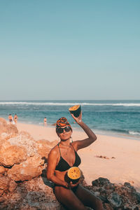 Man wearing sunglasses on rock at beach against sky