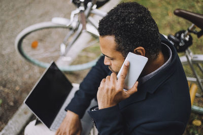 High angle view of male entrepreneur talking on smart phone while sitting with laptop in park