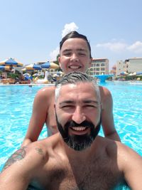 Portrait of smiling father with son enjoying in swimming pool against sky during sunny day