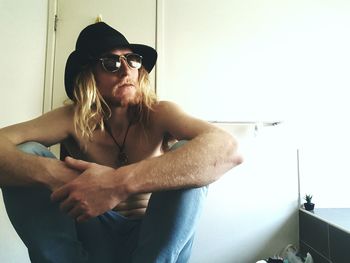 Shirtless young man wearing hat and sunglasses sitting against wall