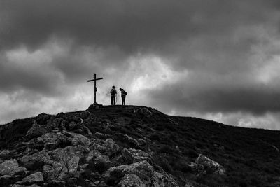 Low angle view of hikers standing by cross on mountain against storm clouds