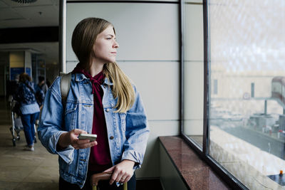 Young woman with smart phone looking out of window at airport