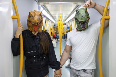 Male and female friends holding hands while wearing dinosaur mask in train