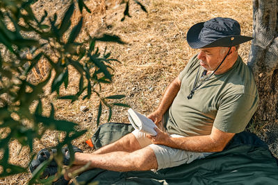 Bearded man tourist relaxing after hiking under the tree with book, reading novel in autumn park.