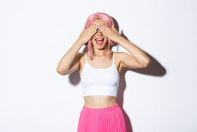Young smiling woman covering eyes standing against wall