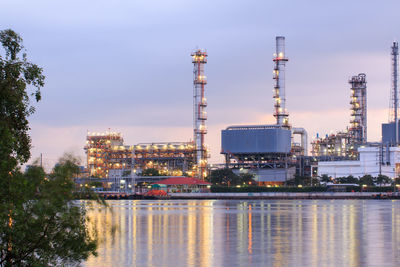 Illuminated factory by river against sky in city