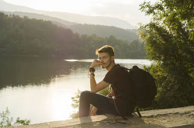 Portrait of man with backpack sitting by lake during sunny day