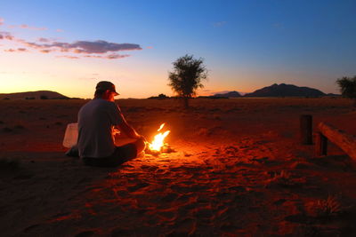Rear view of man sitting by campfire against sky