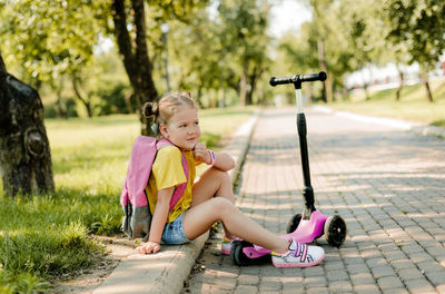 A thoughtful schoolgirl girl is sitting on the sidewalk in the park with a backpack on her shoulders